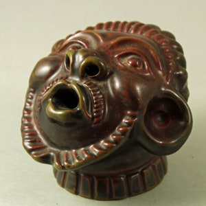 Peut inclure: Une sculpture en céramique marron détaillée d'un visage avec une bouche ouverte et des traits détaillés. La sculpture a une teinte brun rougeâtre avec des accents plus foncés et une bordure décorative autour du visage.
