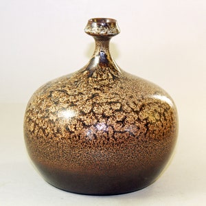 Może przedstawiać: Brązowy ceramiczny wazon o pękatym korpusie i wąskiej szyi. Wazon ma nakrapianą, teksturowaną powierzchnię z ciemnobrązowymi i beżowymi odcieniami. Podstawa jest w ciemniejszym odcieniu brązu.