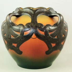 May include: A decorative art glass vase with a bulbous shape. The vase features a gradient of colors, transitioning from green at the base to orange and yellow at the top. Dark brown, ornate metalwork frames the top, adding a contrasting texture and design.
