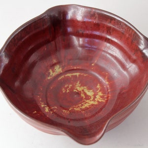 Puede incluir: Un cuenco de cerámica roja hecho a mano con un borde ondulado. El cuenco tiene un acabado brillante con variaciones de color, incluyendo rojo oscuro y toques dorados. El interior presenta círculos concéntricos y una superficie lisa.