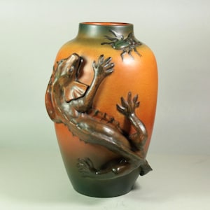 May include: A ceramic vase with a gradient of colors, transitioning from green at the base to orange in the middle and back to green at the top. A detailed brown lizard and insect are sculpted on the vase's surface.