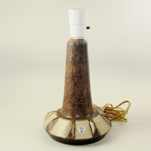 Puede incluir: Lámpara de mesa vintage con base de cerámica marrón y beige moteado. La lámpara tiene un casquillo blanco y un cable eléctrico marrón. La base presenta un patrón decorativo.