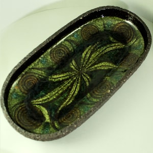 May include: A large oval ceramic serving dish with a dark brown rim and a green and brown geometric design. The design features a central starburst pattern surrounded by swirling circles.