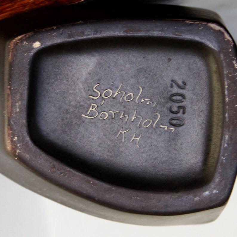 May include: Close-up of the bottom of a dark brown ceramic vase with the text "Sohlom Bornholm KH 2050" inscribed on the bottom.