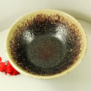 May include: A ceramic bowl with a speckled, dark interior and a light beige rim. The rim has a brown, speckled pattern that fades into the dark interior. The bowl is likely used for serving food.