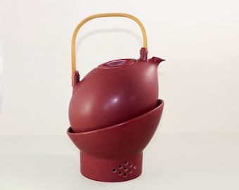 May include: A burgundy ceramic teapot with a wooden handle and a matching burgundy ceramic stand with a strainer. The teapot has a round body and a spout.