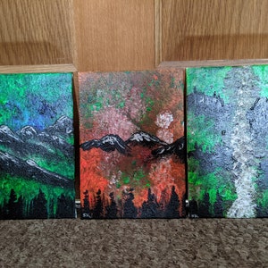 Może przedstawiać: Trzy małe obrazy na płótnie z motywem górskiego krajobrazu. Pierwszy obraz jest zielony, niebieski i biały z czarną sylwetką drzew na dole. Drugi obraz jest czerwony, pomarańczowy i brązowy z czarną sylwetką drzew na dole. Trzeci obraz jest zielony, biały i szary z czarną sylwetką drzew na dole.