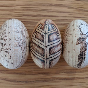 May include: Three wooden eggs with different designs burned into the surface. The first egg has a tree with leaves and raindrops. The second egg has a geometric design. The third egg has a deer with antlers.