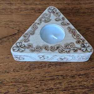 May include: A white triangular candle holder with a carved design of shamrocks and swirls. A small white tea light candle sits inside the holder.