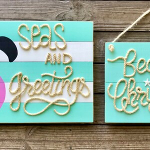 May include: Two wooden signs with a beachy Christmas theme. One sign features a pink flamingo wearing a Santa hat with the text "Seas and Greetings" in rope. The other sign says "Beachy Christmas".