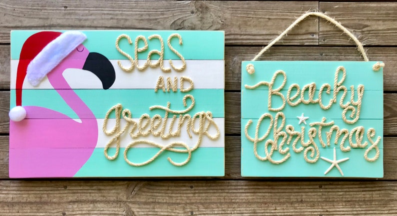May include: Two wooden signs with a beachy Christmas theme. One sign features a pink flamingo wearing a Santa hat with the text "Seas and Greetings" in rope. The other sign says "Beachy Christmas".