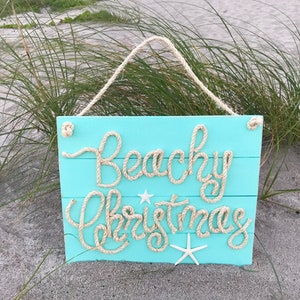 May include: A turquoise wooden sign with the words "Beachy Christmas" spelled out in rope. The sign has a rope hanger and is decorated with starfish. The sign is displayed outdoors.