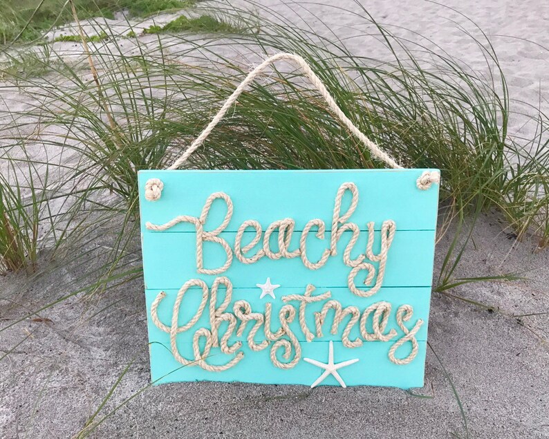 May include: A turquoise wooden sign with the words "Beachy Christmas" spelled out in rope. The sign has a rope hanger and is decorated with starfish. The sign is displayed outdoors.