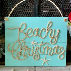 May include: A turquoise wooden sign with the words "Beachy Christmas" spelled out in rope. The sign is decorated with starfish and has a rope handle. Several starfish are placed below the sign.