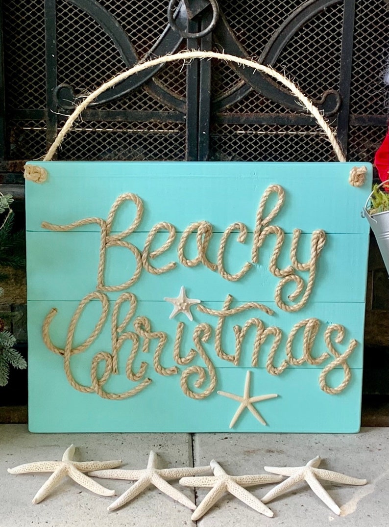 May include: A turquoise wooden sign with the words "Beachy Christmas" spelled out in rope. The sign is decorated with starfish and has a rope handle. Several starfish are placed below the sign.