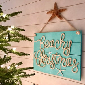 May include: A turquoise wooden sign with the words "Beachy Christmas" in rope lettering. The sign is decorated with starfish and hung by a rope. A natural starfish is at the top. A Christmas tree is in the background.