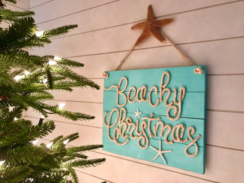 May include: A turquoise wooden sign with the words "Beachy Christmas" in rope lettering. The sign is decorated with starfish and hung by a rope. A natural starfish is at the top. A Christmas tree is in the background.