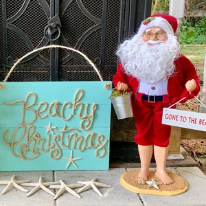 May include: A turquoise wooden sign with the words "Beachy Christmas" in rope lettering, starfish, and a Santa Claus figurine in a red suit holding a sign that says "Gone to the Beach".