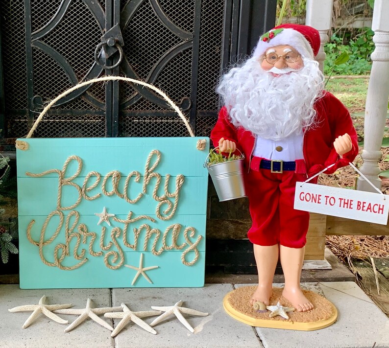 May include: A turquoise wooden sign with the words "Beachy Christmas" in rope lettering, starfish, and a Santa Claus figurine in a red suit holding a sign that says "Gone to the Beach".