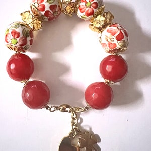 Könnte beinhalten: Ein Armband mit abwechselnd roten und floralen Perlen. Die floralen Perlen sind weiß mit roten Blumen und goldenen Details. Ein goldener Anhänger mit einem Herz und einem vierblättrigen Kleeblatt hängt am Verschluss.