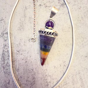 Op de afbeelding: Een zilveren ketting met een hanger met een paarse amethistsfeer en een veelkleurig, kegelvormig kristal. Het kristal toont kleurstroken, waaronder paars, blauw, groen, geel en rood. De ketting ligt op een lichtgrijze ondergrond.
