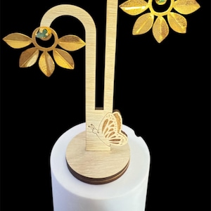 Puede incluir: Pendientes dorados en forma de hoja con un pequeño centro de piedra verde, exhibidos en un expositor de joyería de madera. El expositor presenta un recorte de mariposa y está colocado sobre una base cilíndrica blanca sobre un fondo negro.