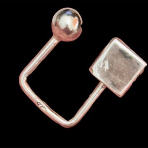Puede incluir: Un pendiente plateado con un diseño geométrico. El pendiente presenta una pequeña esfera en la parte superior, una forma cuadrada en la parte inferior y una barra doblada que conecta las dos formas. El pendiente está sobre un fondo negro.