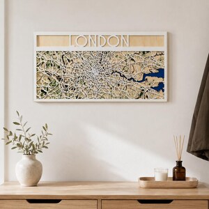 Könnte beinhalten: Ein rechteckiges Wandbild mit einer detaillierten, lasergeschnittenen Karte von London. Die Karte zeigt komplexe weiße Straßennetze über einer beigen und grünen Landschaft mit einem blauen Flussabschnitt. Das Wort LONDON ist oben in Weiß ausgeschnitten.