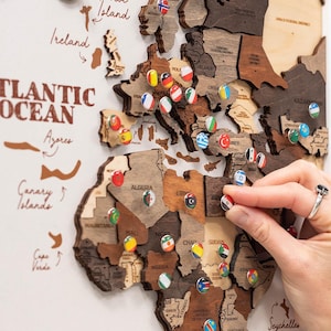 May include: A wooden wall map featuring laser-cut continents in varying shades of brown, with small circular pins displaying national flags placed on specific countries. The text ATLANTIC OCEAN is printed in brown on the light background.