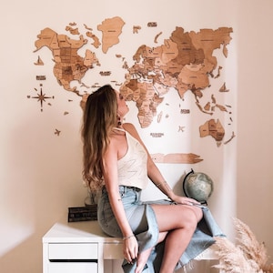 May include: A detailed wooden world map wall decoration, with country outlines. The map is mounted on a light wall. A person sits on a white desk, with a globe and plants nearby. The map is a natural wood colour.