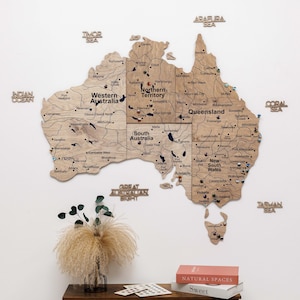 May include: A detailed wooden map of Australia, showing states and cities, mounted on a white wall. Pins mark locations. A table below displays books and decorative items, including a vase of dried flowers.