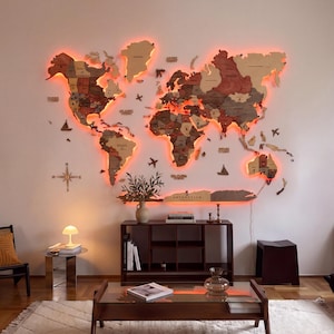 Peut inclure: Une grande carte du monde murale en bois multicouche, composée de différentes nuances de brun et de beige, illuminée par un rétroéclairage LED orange vif. La carte comprend des étiquettes de pays, une rose des vents et des icônes en bois.