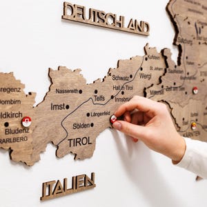 May include: A laser-cut wooden wall map of Austria featuring the region of Tirol with engraved city names like Innsbruck, Telfs, and Sölden. Small circular pins with red and white emblems are placed on specific locations across the map surface.