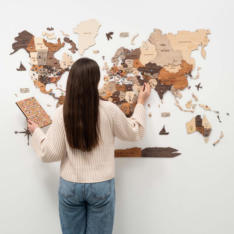 Puede incluir: Un mapa del mundo de madera con contornos de pa&iacute;ses y alfileres, montado en una pared blanca. El mapa est&aacute; hecho de varias tonalidades de madera marr&oacute;n. Se ve a una persona colocando alfileres en el mapa, sosteniendo una peque&ntilde;a tabla con alfileres adicionales.