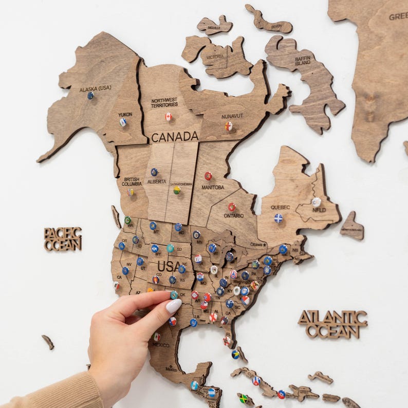 Puede incluir: Mapa del mundo de madera que muestra Am&eacute;rica del Norte, con alfileres que marcan ubicaciones en los EE. UU. y Canad&aacute;. El mapa es de madera marr&oacute;n claro e incluye el texto "PACIFIC OCEAN" y "ATLANTIC OCEAN". Una mano coloca un alfiler en el mapa.
