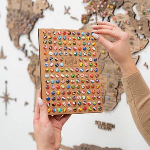 May include: A close-up of a brown wooden board with a grid of colourful round pins. Each pin features a different country's flag. The board is labelled "Country Flags" and "Enjoy the Wood".