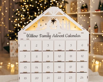 May include: A white wooden Advent calendar shaped like a house, with 24 numbered drawers. The top features a cutout nativity scene and the text "Willow Family Advent Calendar". The calendar is illuminated from within, creating a warm glow.