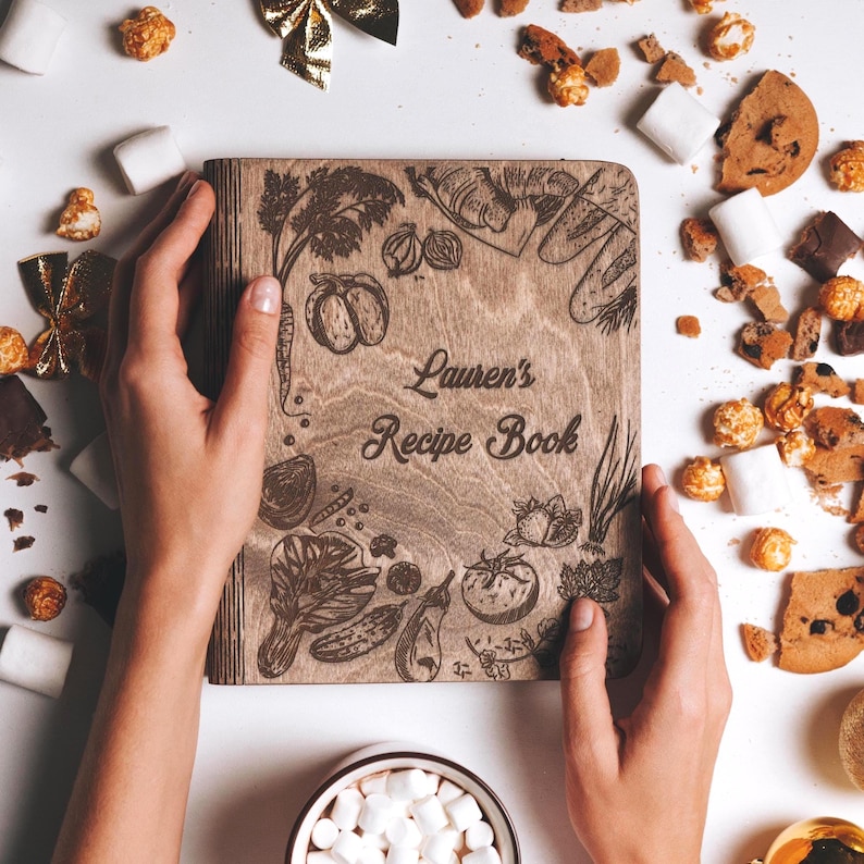 K&ouml;nnte beinhalten: Ein Rezeptbuch aus Holz mit einem braunen Holzdeckel und einem lasergravierten Design aus Gem&uuml;se und dem Text "Lauren's Recipe Book".