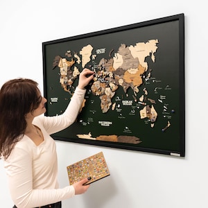 May include: A wooden world map in a black frame, mounted on a wall. The map features different wood tones and pins. A person is placing a pin on the map, holding a box of pins. The background is a plain white wall.