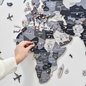 May include: A multi-layered wooden world map wall art featuring various shades of gray and black. The map includes laser-cut country names, borders, and decorative airplane icons. Small circular pins with national flags are placed across the map.