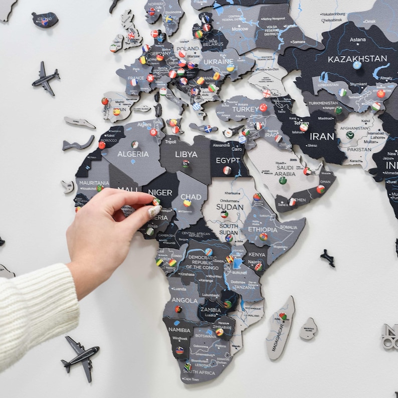 May include: A multi-layered wooden world map wall art featuring various shades of gray and black. The map includes laser-cut country names, borders, and decorative airplane icons. Small circular pins with national flags are placed across the map.