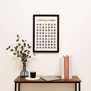 May include: A framed print titled "NATIONAL PARKS" with a grid of colorful park icons. The black frame hangs on a white wall above a wooden table. A vase with greenery, a candle, and books are on the table.