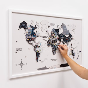 Peut inclure: Une carte du monde encadrée avec un cadre blanc, présentant un schéma de couleurs noir, gris et blanc. La carte est ornée d'épingles colorées, indiquant les destinations de voyage. La rose des vents et les noms des océans sont visibles.