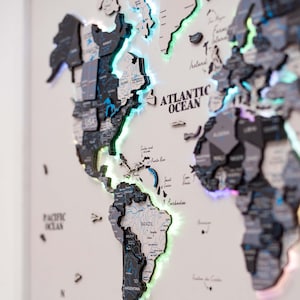 Op de afbeelding: Een gedetailleerde, driedimensionale wereldkaart met verlichte randen. De kaart heeft een gelaagd ontwerp met individuele landuitsparingen in grijstinten en zwart. De Atlantische Oceaan is gemarkeerd. De kaart is op een wit oppervlak gemonteerd.