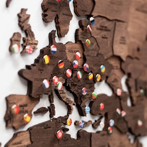 May include: A wooden map of Europe with country outlines and pins. The map is made of dark brown wood and has pins with various country flags. The map is mounted on a white surface.