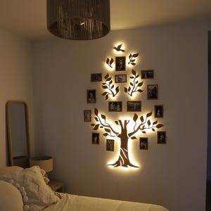 Puede incluir: Una pieza de arte de pared de madera con forma de &aacute;rbol con ramas y hojas. El &aacute;rbol est&aacute; iluminado con luces LED blancas c&aacute;lidas. Hay peque&ntilde;os marcos de fotos colgados de las ramas. El &aacute;rbol est&aacute; montado en una pared blanca en un dormitorio.