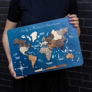 Puede incluir: Un mapa del mundo rectangular de madera para pared con un fondo azul oscuro texturizado. Los continentes están hechos de madera en capas con tonos marrones variados. Incluye el texto Emily & Michael's Adventures, nombres de océanos y una rosa de los vientos.