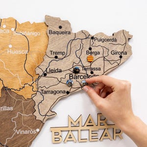 Może przedstawiać: Drewniana mapa Katalonii z nazwami miast i znacznikami pinezek. Mapa jest jasnobrązowa z ciemnobrązowymi liniami wskazującymi drogi. Słowo "MAR BALEAR" znajduje się na dole. Ręka umieszcza pinezkę na mapie.