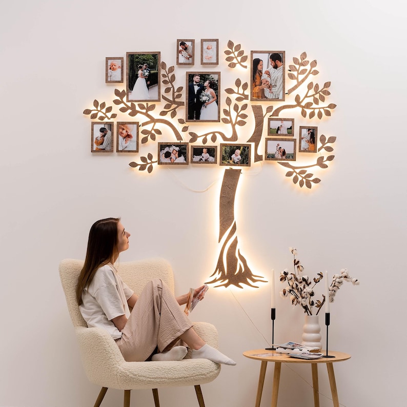 May include: A wooden wall decor in the shape of a tree with lights and multiple framed photos of people. The tree is lit up with warm white lights. A person is sitting in a white armchair in front of the tree.