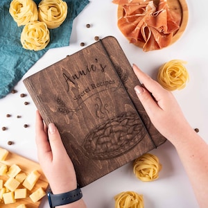 K&ouml;nnte beinhalten: Ein dunkelbraunes Rezeptbuch aus Holz mit dem Text "Annie's Secret Recipes" auf dem Einband. Das Buch hat ein dekoratives Design mit einer Torte und einem Strudel aus Dampf.